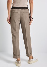 Personne portant un pantalon beige sur mesure avec des ourlets retroussés et des baskets blanches, vue de dos contre un fond uni.
