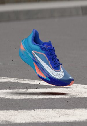 Chaussure de course Nike Zoom Fly bleue et orange flottant au-dessus d'une route asphaltée avec des lignes blanches peintes et de petits débris.
