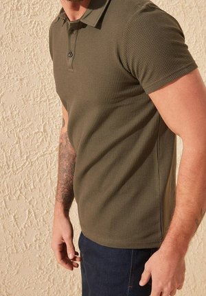 Uomo che indossa una polo a maniche corte verde oliva con trama e pantaloni blu scuro, in piedi davanti a un muro beige texturizzato.
