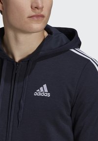 Sudadera con cremallera en color navy, con capucha ajustable, logo blanco de Adidas y tres franjas blancas en cada hombro. Textura de tela suave.
