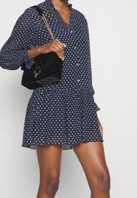 Kvinna i en långärmad klänning med marinblåa polka prickar som håller en svart sammetshandväska med en guldkedjeaxelrem.