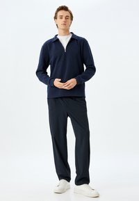 Marineblauwe pullover met een ritskraag, gestructureerde stof en ribgebreide zoom. Gecombineerd met donkere broek en witte sneakers, staand tegen een lichte achtergrond.
