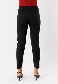 Donna che indossa pantaloni neri slim-fit, top rosso e sandali neri con tacco, in piedi contro uno sfondo bianco semplice, vista da dietro.