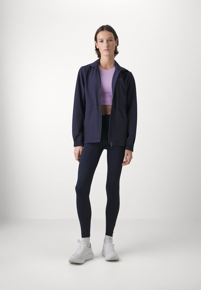 Navy zip-up jacket over a purple crop top, paired with navy leggings and white sneakers. The jacket features elastic cuffs and pockets.
