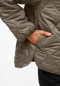 Main dans la poche d'une veste grise matelassée avec des coutures courbes, portée sur un pantalon noir contre un fond uni.