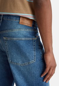 Niebieskie dżinsy o dopasowanym kroju, z brązową skórzaną metką, szwami na tylnych kieszeniach i gładką fakturą.