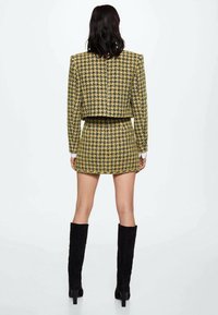 Keltainen ja musta houndstooth-kuvioitu lyhyt takki yhdistettynä yhteensopivaan minihameeseen ja reiteen yltäviin mustiin korkokenkiin. Sujuva kangas.