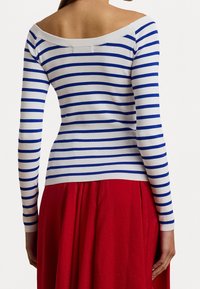 Pull rayé bleu et blanc à col bateau ; coupe ajustée en maille avec manches longues, texture douce, porté avec une jupe rouge fluide.