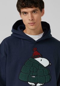 Marineblauwe hoodie met een gestructureerd, groen gewatteerd ontwerp en een geborduurd figuur met een rode hoed.