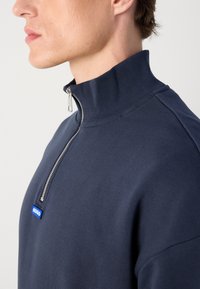 Sudadera de media cremallera en azul marino, fabricada en un tejido suave con cuello acanalado. Presenta una cremallera plateada y un logo azul de "HUGO" en la parte frontal.
