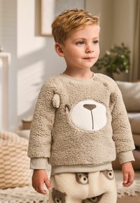 Beige, fluffy sweater with a bear face design, round white patch, and button detail. Paired with patterned fleece pants featuring bear faces.