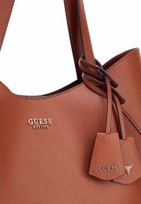Sac fourre-tout en faux cuir marron avec une finition texturée, présentant des accents de logo en métal argenté et deux petites étiquettes attachées aux poignées.
