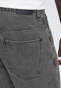 Blugi gri din denim cu buzunar în spate, prevăzut cu un detaliu subtil de cusătură și o patch piele neagră deasupra taliei. Textură netedă vizibilă.