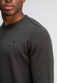 Dunkelgrauer Pullover aus einem weichen Material, mit einem gerippten Halsausschnitt und einem kleinen grünen Logo-Patch auf der Brust.