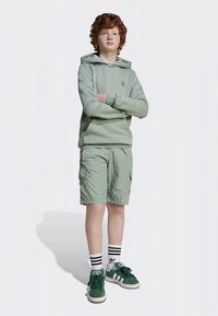 Light green hoodie with kangaroo pocket, paired with light green cargo shorts. Wears green sneakers with white accents and striped socks.
