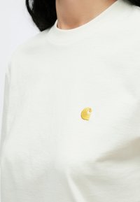 Mujer con una camiseta blanca lisa con un pequeño logo bordado en amarillo en el lado izquierdo del pecho, recortada justo debajo de la barbilla.