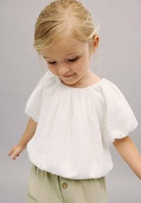 Blusa bianca in cotone a maniche corte con scollo arricciato e dettagli plissettati. Abbinata a pantaloni beige chiaro con un solo bottone.