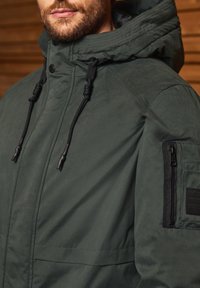 Grön jacka med en dragkedjeficka på vänster arm, justerbar huva med snoddar och en matt yta.