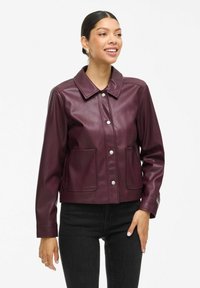 Veste en simili cuir bordeaux avec un col, fermeture à boutons-pression, deux poches avant et manches longues, portée avec un jean noir.