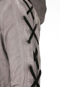 Grau Denim-Jacke mit schwarzen Spitzenakzenten, die durch Metallösen entlang des Ärmels gefädelt sind. Der Stoff hat eine weiche, strukturierte Oberfläche.