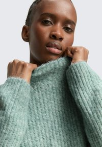 Mintgrüner, grob gestrickter Pullover mit hohem Kragen und gerippter Textur. Der Stoff wirkt weich, mit sichtbaren Fasern und einer lockeren Passform.