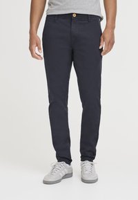 Tamnoplave chinos hlače sa slim fit krojem, prednjim džepovima, jednim dugmetom za zatvaranje i suptilnom teksturom. Uparene s svijetlosivim tenisicama.