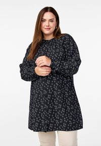 Blouse noire avec un motif en diamants blancs. Le matériau est doux avec une texture légèrement brillante, un col rond et une fermeture à boutons.