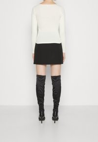 Langärmliges, cremefarbenes Oberteil, kombiniert mit einem schwarzen Minirock. Schwarze satinierte Overkneestiefel mit spitzem Zehenbereich und Stiletto-Absatz. Schlichtes und enganliegendes Design.