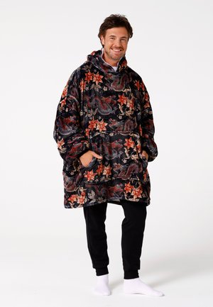 Zwart oversized velvet poncho met een bloemen- en drakenpatroon in rood en goud, met een capuchon en zakken aan de voorkant, gedragen over een zwarte broek.