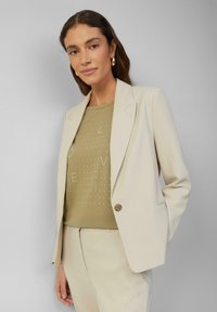 Blazer beige ajusté avec revers en pointe, fermeture à un bouton, et un pantalon assorti. T-shirt vert olive avec un motif à pois.