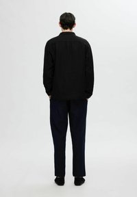 Juodas, ilgomis rankovėmis marškiniai, galinčiais pasirodyti kaip linas, derinami su tamsiomis, laisvos talpos kelnėmis. Pateikiama iš nugaros.