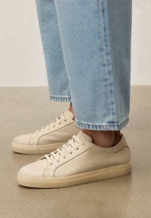 Osoba stojící na béžové podlaze, oblečená v světle modrých džínách a krémových kožených teniskách s nízkým střihem.