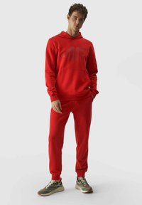 Sudadera roja con pantalones jogger a juego, hechos de una tela suave. La sudadera cuenta con un bolsillo delantero y un gran logotipo. Las zapatillas son grises con acentos naranjas.