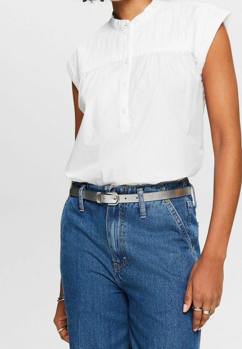 Femme portant un chemisier blanc sans manches avec des boutons, rentré dans un jean bleu taille haute, ceinturé d'une ceinture argentée et portant des bagues aux doigts.