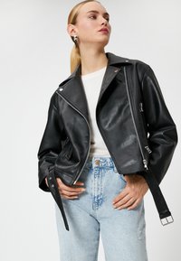 Sort lædermotorcykeljakke med sølvhardware, lynlåslommer og en tætsiddende pasform. Båret over en hvid skjorte og lyse denimjeans.