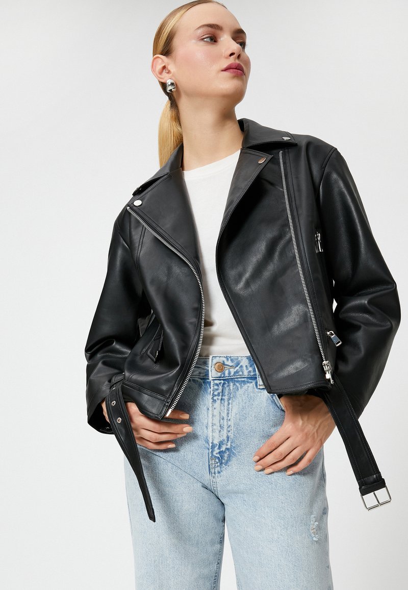 Sort lædermotorcykeljakke med sølvhardware, lynlåslommer og en tætsiddende pasform. Båret over en hvid skjorte og lyse denimjeans.