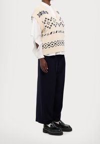 Gilet beige en tricot avec col en V et motif bleu marine, porté sur une chemise blanche aux manches larges, associé à un pantalon bleu marine et des chaussures noires.