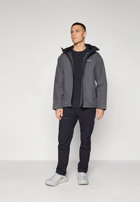 Casaco impermeável cinza com capuz, fecho de correr frontal e punhos elásticos, combinado com calças pretas e ténis cinza claro. Design simples e funcional.