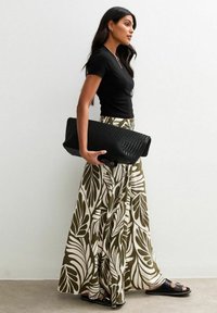 Black fitted t-shirt, olive-green skirt with white leaf pattern, large black textured clutch, and black flat sandals, standing against a wall.