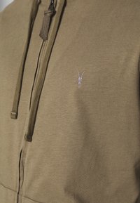 Sudadera con capucha verde oliva con cremallera, hecha de algodón suave, con capucha ajustable mediante cordón y un pequeño logo de calavera rosa en el lado izquierdo del pecho.