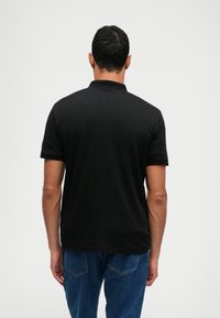 Black polo shirt with short sleeves, textured collar, and a smooth fabric finish. Worn with blue denim jeans, viewed from the back.