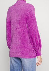 Fuzzy lila pullover med hög hals, ballongsärmar och avslappnad passform. Tillverkad av mjukt, texturerat material. Kombineras med ljusblå jeans.