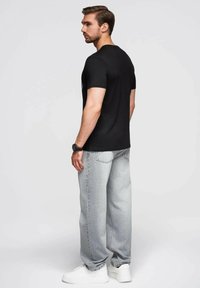 Černé tričko s krátkým rukávem, šedé volné džíny a bílé tenisky. Model stojí zády, ukazuje jednoduchý design a uvolněný střih.