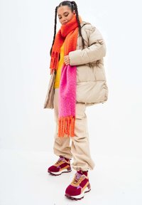 Giacca a vento beige, sciarpa con gradiente arancione e rosa, maglione giallo, pantaloni beige e sneaker bordeaux con accenti viola e gialli.