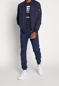 Granatowa kurtka bombowca z zamkiem błyskawicznym, granatowy sweter z białym logo, granatowe spodnie dresowe, a także białe sneakersy na jednolitym tle.