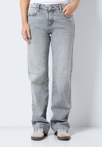 Personne portant un jean droit gris clair avec les mains dans les poches et des baskets blanches à semelles marron, debout devant un arrière-plan uni.