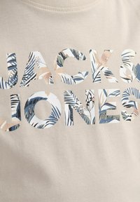 Bež majica s natpisom "JACK & JONES" u izrezanim slovima ispunjenim cvjetnim uzorkom u zelenim, ružičastim i plavim tonovima. Pamuk.