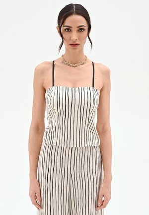 Kobieta ubrana w białą i czarną, pionowo paskowaną, bezrękawną bluzkę oraz pasujące szerokie spodnie z nogawkami o luźnym kroju, z naszyjnikiem ze złotego łańcucha.