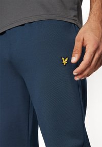 Navy blue athletic pants with a smooth texture, featuring a small yellow embroidered logo of an eagle on the thigh area.