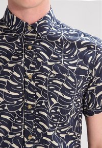 Camicia a maniche corte con fondo navy e motivo a foglie bianche, caratterizzata da un colletto classico, bottoni e una tasca sul petto.